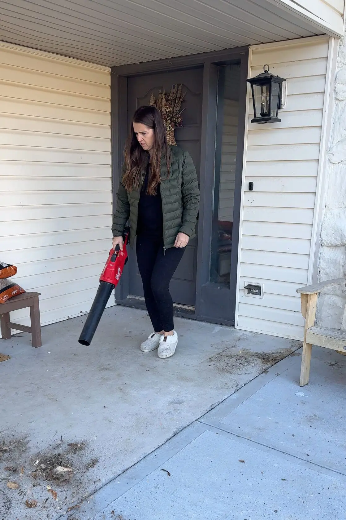 A woman using a red leaf blower to clean dirt and leaves from a front porch, dressed in a green puffer jacket and casual black leggings, standing in front of a gray door with white siding.